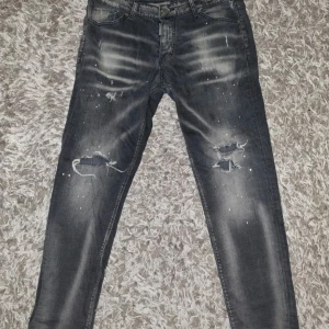 Svarta Dsquared2 jeans med slitningar - Säljer ett par svarta jeans från Dsquared2 med coola slitningar och färgstänk på framsidan. Jeansen har en snygg tvättad look, raka ben och klassisk femficksdesign. Perfekta för dig som gillar streetstyle och vill sticka ut lite extra.