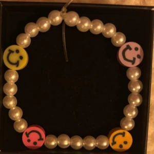 Pärlarmband med smileys i färg - Ett pärlarmband med vita pärlor och fyra färgglada smiley-berlocker i gult, rosa och lila. Armbandet har en lekfull och trendig stil som passar perfekt för dig som gillar färg och glada detaljer.