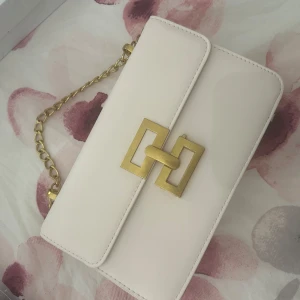 Vit axelväska med guldkedja - Stilren vit axelväska i skinn med guldfärgad kedja och snyggt geometriskt spänne framtill. Väskan har ett rymligt fack och elegant design som lyfter vilken outfit som helst. Perfekt storlek för det viktigaste och en riktig statement piece.