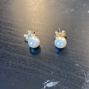 Guldörhängen med pärlor och rosett - Supersöta örhängen med guldfärgade rosetter och stora vita pärlor. Perfekt om du vill ha en elegant och gullig vibe. Kommer med en blå smyckespåse. Säljs som ett par. Köpta på ur & Penn, vet inte om de är nickelfria 