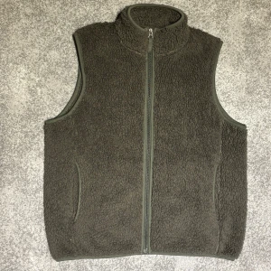Mörkgrå fleeceväst från Uniqlo - Mysig mörkgrå fleeceväst från Uniqlo i storlek XS. Västen har hög krage, hel dragkedja framtill och två fickor på sidorna. Tillverkad i 100% polyester med en fluffig och varm känsla, perfekt att slänga på sig över en hoodie eller tröja.