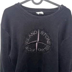 Svart sweatshirt från Stone Island - Svart sweatshirt från Stone Island med stort broderat kompassmärke på bröstet och klassisk patch på ärmen. Tröjan har rund hals, ribbade muddar och är tillverkad i mjuk bomull. Perfekt för dig som gillar streetwear och vill ha en clean look.
