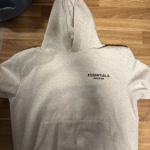 Essentials Fear of God grå hoodie - Säljer en ljusgrå hoodie från Essentials Fear of God med svart logga på bröstet och ryggen. Tröjan har huva, känguruficka och är tillverkad i mjukt bomullsmaterial. Perfekt för en chill och stilren look.