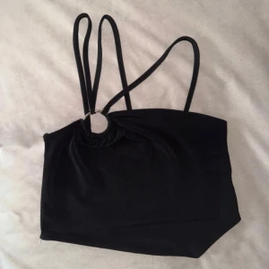 Svart topp med ring och smala band - Snygg svart topp med tre unika smala axelband och en silverfärgad ringdetalj framtill. Toppen har en asymmetrisk nederkant framtill. Perfekt för att skapa en trendig look på alla höstens fester. Väldigt fint skick på denna topp!