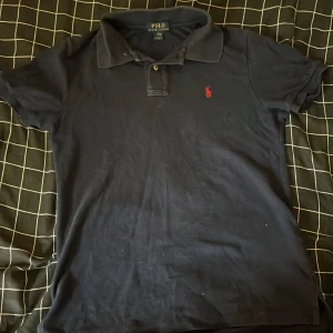 Mörkblå pikétröja från Polo Ralph Lauren - Snygg mörkblå pikétröja från Polo Ralph Lauren med klassisk krage och två knappar. Tröjan har korta ärmar och är prydd med det ikoniska röda broderade logotypen på bröstet. Tillverkad i mjuk bomull för en skön känsla.