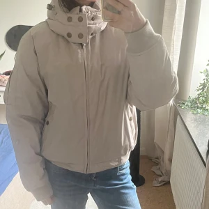Beige dunjacka från Wolf Wear - Snygg beige dunjacka från Wolf Wear med hög krage, stora knappar och dragkedja. Jackan har en pufferstil, ribbade muddar och praktiska fickor. Fylld med dun och fjäder, yttertyg i polyester och bomull. Perfekt för kalla dagar. Uppskattas vara i strl S/M. Avtagbar luva
