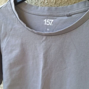 Grå basic t-shirt från 157 XS - Enkel och stilren grå t-shirt från 157 i storlek XS. T-shirten har rund halsringning och korta ärmar, perfekt för en clean och avslappnad look. Tillverkad i mjuk bomull som känns skön mot huden.