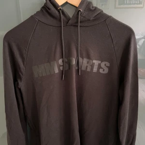 Svart hoodie från MM Sports - Svart hoodie från MM Sports med tryckt logga över bröstet. Tröjan har huva med snörning och långa ärmar. Perfekt för träning eller chill dagar. Mjuk och bekväm med klassisk passform.