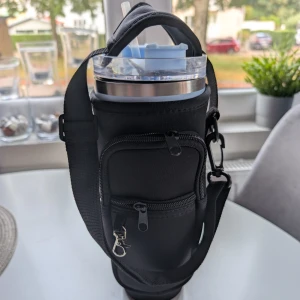 Svart temos väska med temos - Praktisk set med blå  termosmugg svart flaskväska med justerbar axelrem och två dragkedjefickor framtill. Plus extra sugrörslock, så du slipper spill. Perfekt för att bära vattenflaska eller termos, med plats för småprylar i fickorna. Väskan är tillverkad i mjukt syntetmaterial och har en karbinhake för extra fästmöjligheter.