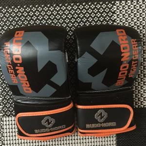 Svarta Budo-Nord fight handskar L - Säljer ett par svarta fight handskar från Budo-Nord med grått och orange tryck. De har breda kardborreband med orange kant och är märkta med 'Twin Layer Safety'. Perfekta för kampsport eller träning. Säckhandskar som knappt är använda!