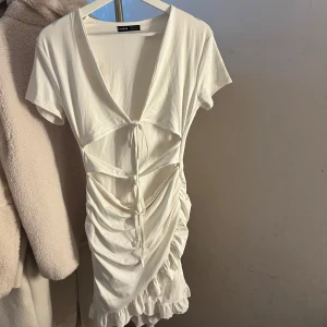 Kort omlottklänning från SHEIN - Supersnygg kort klänning från SHEIN med omlott-design, snörning framtill och volangkant nedtill. Finns i tre färger: vit, ljusblå och orange. Klänningen har v-ringning, korta ärmar och är gjord i mjukt polyester-material. Perfekt för dig som vill sticka ut med coola detaljer.