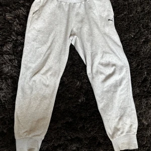 Grå mjukisbyxor från Puma - Snygga grå mjukisbyxor från Puma med elastisk midja och muddar vid bensluten. Byxorna har en klassisk passform och är tillverkade i mjuk bomullsblandning. Puma-loggan i svart finns på ena benet. Perfekta för chill eller träning. Defekt syns på sista bilden.