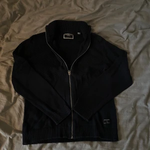 Mörkblå kofta från Jack & Jones - Säljer en snygg mörkblå full zip från Jack & Jones med dragkedja framtill. Perfekt för kyliga dagar med sin stickade design och höga krage. Passar både till vardags och lite finare tillfällen. Skön och stilren!