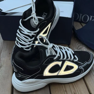 Dior B30 skor - Säljer ett par Dior B30 sneakers i svart med vita och reflekterande loggor. Skorna har tydliga CD-loggor på sidorna, vit sula och vita snören. Ovandelen är i mesh och syntet, vilket ger en sportig och modern vibe. Perfekta för dig som vill sticka ut med exklusiva sneakers. Extra snören, tags, box och alla papper medföljer såklart.