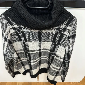 Svartvit rutig polotröja - Snygg svartvit polotröja med rutigt mönster och bred, ribbad polokrage. Tröjan är stickad och har en loose passform med långa ärmar. 