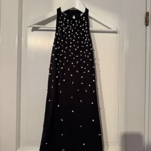 Svart glittrig kortklänning med pärlor - Supersnygg svart kortklänning från Zara med glittriga stenar och pärlor över hela framsidan. Klänningen har hög hals och är ärmlös, vilket ger en cool och elegant vibe. Perfekt för dig som vill sticka ut med lite extra bling.