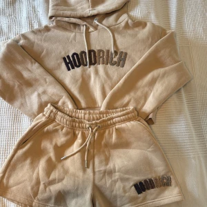 Beige Hoodrich shorts med snörning XS - Snygga beige set från Hoodrich i mjuk bomull med elastisk midja och snörning. Broderad Hoodrich-logga i svart och brunt på benet och på hoodie. Perfekt för chill dagar och streetwear-vibe. Modellen är loose fit och har praktiska sidofickor både på shortsen och på själva hoodie 