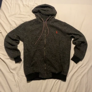 Mörkgrå ralph lauren hoodie  - Mörkgrå hoodie från Polo Ralph Lauren med dragkedja, huva och snörning. Tröjan har ett rött broderat logomärke på bröstet och svarta muddar vid ärmslut och nederkant. Perfekt för dig som gillar klassisk streetstyle.