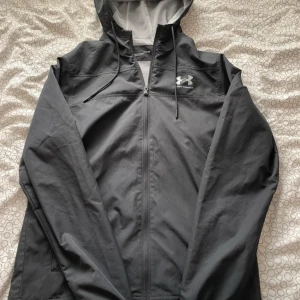 Svart vindjacka från Under Armour S - Svart vindjacka från Under Armour med huva och dragkedja framtill. Jackan har meshfoder, snörning i huvan och logga på bröstet. Lätt och sportig modell med loose fit, perfekt för utomhusaktiviteter. Pris kan diskuteras!