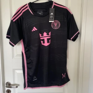 Inter Miami CF svart fotbollströja Adidas - Svart Inter Miami CF fotbollströja från Adidas med rosa detaljer och klubbmärke på bröstet. Tröjan har korta ärmar, rund hals och är tillverkad i lätt och ventilerande polyester. Snygga rosa ränder på axlarna och tryck på både fram- och baksida.