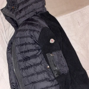 Svart Moncler pufferjacka med stickade ärmar - Snygg svart pufferjacka från Moncler med stickade ärmar och detaljer. Jackan har dragkedja framtill, hög krage och Moncler-logga på ärmen. Perfekt för dig som vill ha en stilren och varm jacka med exklusiv känsla.