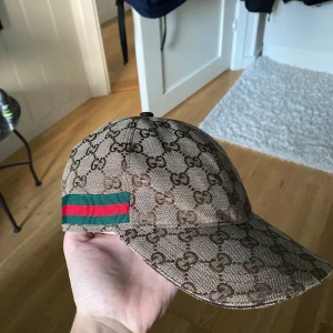 Snygg beige keps - Säljer en snygg keps  i beige och brunt med klassiskt GG-monogram över hela kepsen. Kepsen har en grön och röd rand på sidan och är tillverkad i bomullsmix med justerbar rem baktill. Perfekt för dig som vill sticka ut med en lyxig accessoar.