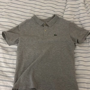 Grå pikétröja från Lacoste - Snygg grå pikétröja från Lacoste med klassisk krage och två knappar framtill. Ikoniska gröna krokodilloggan på bröstet. Tillverkad i mjuk bomull, perfekt för en clean och stilren look. Liten i storlek❗️använd ett få tal gånger. 9/10 kvalite