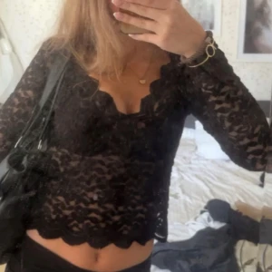 Svart spetsblus från Zara - Säljer en svart långärmad spetsblus från Zara med v-ringning och vågig kant nertill. Blusen är genombruten och har en feminin, croppad passform som ger en snygg siluett. Perfekt för dig som vill sticka ut med en elegant och trendig topp.