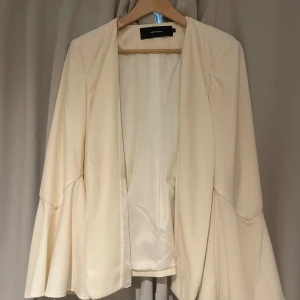 Beige kavaj med utsvängda ärmar Vero Moda - Stilren beige kavaj från Vero Moda med snygga utsvängda ärmar. Jackan har öppen front utan knäppning och en figurnära passform. Perfekt för dig som vill ha en trendig och elegant look med en twist.