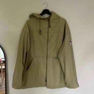 Beige/grön vindjacka med huva  - Stilren beige/grön vindjacka med huva och dragkedja framtill. Jackan har två stora fickor och en patch på ärmen. Perfekt för blåsiga dagar och enkel att matcha med det mesta.