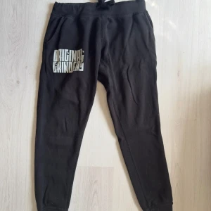 Svarta mjukisbyxor Original Grinders - Svarta mjukisbyxor från Original Grinders med tryck på vänster lår och båda vaderna. Byxorna har resår och snörning i midjan, samt en bakficka. Perfekta för chill eller träning, tillverkade i mjuk bomull.