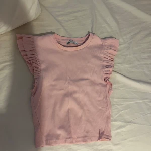 Rosa ribbad topp med volangärmar Zara - Supersöt ljusrosa ribbad topp från Zara med volangdetaljer på axlarna. Toppen är ärmlös och har en rund halsringning. Perfekt för dig som gillar en feminin och trendig stil.