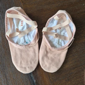 Ballerinaskor i ljusrosa från Bloch - Ljusrosa ballerinaskor från Bloch med korsade resårband över foten. Skorna är i bomullstyg med mockadetaljer under sulan och har en klassisk rund tå. Perfekta för dans eller balettträning. Storleken är 4 1/2 C motsvarande 35,5