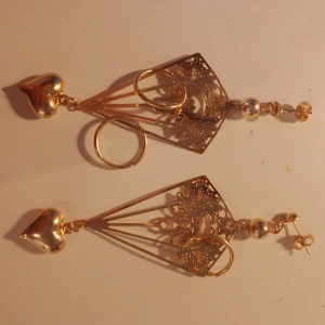 Guldiga örhängen  - Unika guldiga örhängen med stora hjärtformade detaljer upptill, dekorativa cirklar och ett utskuret, ornamenterat triangulärt mönster. Perfekta för dig som vill sticka ut med cool design och snygga detaljer.