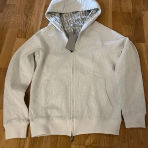 Vit hoodie från Dior med monogramfoder - Snygg vit hoodie från Dior med hel dragkedja och huva. Insidan är fodrad med Diors ikoniska monogrammönster i grått. Tröjan har två stora fickor framtill och ribbade muddar. Perfekt för dig som vill ha en exklusiv och stilren look.