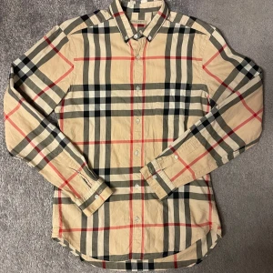 Rutig Burberry Brit skjorta beige - Klassisk rutig skjorta från Burberry Brit i beige med svarta, vita och röda rutor. Skjortan har lång ärm, knappar framtill och button down-krage. Tillverkad i mjuk bomull och har en tidlös, snygg passform som funkar till många stilar.