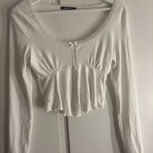 Vit croppad blus med spets från Gina Tricot - Supersöt vit croppad blus från Gina Tricot med långa ärmar, spetsdetaljer längs urringningen och en liten rosett framtill. Säljer den pga att den knappast kommer till användning.