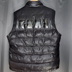 Hugo Boss Dunväst - Snygg svart dunväst från Hugo Boss med diskret logga på bröstet och stor logotyp i blankt material på ryggen. Västen har hög krage, dragkedja framtill och två fickor med tryckknapp. Perfekt för lager-på-lager och streetwear-vibe.