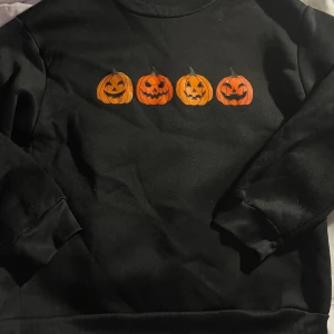 Svart sweatshirt med pumpor, M - Svart sweatshirt i 100% polyester med tryck av fyra orange pumpor med olika ansiktsuttryck på bröstet. Tröjan har rund hals, ribbade muddar vid ärmslut och nederkant samt lång ärm. Perfekt för dig som gillar Halloween-vibes.