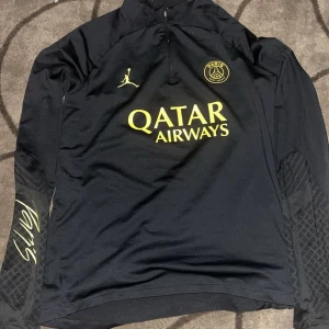 PSG långärmad träningströja Jordan - Snygg svart långärmad PSG träningströja från Jordan med gul logga och detaljer. Tröjan har halv dragkedja, broderad Jumpman och Paris-märke på bröstet samt Qatar Airways-tryck. Ärmarna har diskret mönster och signaturdetalj. Perfekt för fotbollsträning eller streetwear.