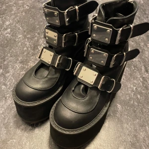 Svarta platåboots med spännen - Riktigt feta svarta boots med extrem platåsula och fyra breda spännen med metallplattor. Skorna är i skinnimitation och har rund tå. Perfekta för dig som vill sticka ut och få extra höjd. 