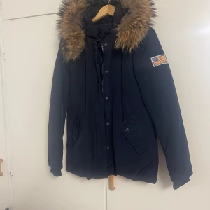 Mörkblå parkas från Svea med fuskpäls - Snygg mörkblå parkas från Svea med stor huva och fuskpäls runt kanten. Jackan har dragkedja och knappar framtill, två fickor med lock och en amerikansk flagga på ärmen. Perfekt för kalla dagar och riktigt skön passform.