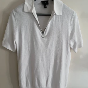Vit piké-tshirt med dragkedja H&M M - Vit piké-tshirt från H&M i slim fit med korta ärmar och krage. Tröjan har en snygg silverfärgad dragkedja framtill med ringdetalj och är gjord i ett strukturerat bomullstyg. Perfekt för en clean och fräsch look.