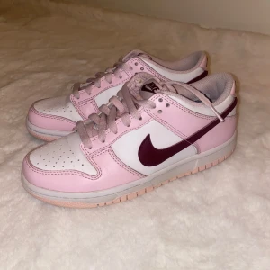 Nike Dunk Low rosa och vita sneakers - Nike Dunk Low sneakers i en fräsch kombo av ljusrosa och vitt med vinröda detaljer och swoosh. Skorna har klassisk snörning, rund tå och platt sula. 