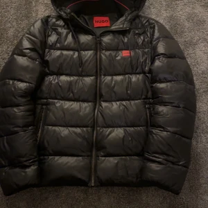 Svart pufferjacka från HUGO - Säljer en svart pufferjacka från HUGO med huva och dragkedja framtill. Jackan har två sidofickor med dragkedja och en röd HUGO-logga på bröstet. Perfekt för kalla dagar och har en stilren look med quiltad design. Nypris 3500kr