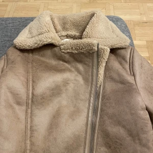 Beige teddyjacka med dragkedja - Säljer en beige teddyjacka från STR med mjukt foder och stor krage. Jackan har en schysst dragkedja framtill och är fodrad med fluffigt teddy-material. Perfekt för kalla dagar och har en snygg, lite boxig passform. Ärmarna är långa och har uppvikta muddar.