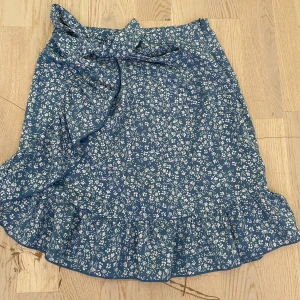 Blommig blå omlottkjol med volang - Supersöt blå kortkjol med vitt blommönster och volangkant. Kjolen har omlottmodell och knyts i midjan med ett band. Perfekt för dig som gillar en romantisk och somrig stil.