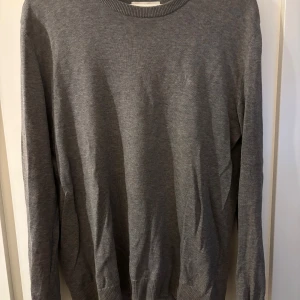 Grå stickad tröja med rund hals - En stilren grå stickad tröja med rund halsringning och långa ärmar. Tröjan har ribbade muddar vid ärmslut och nederkant, perfekt för lager-på-lager. Enkel och klassisk design som passar till det mesta.