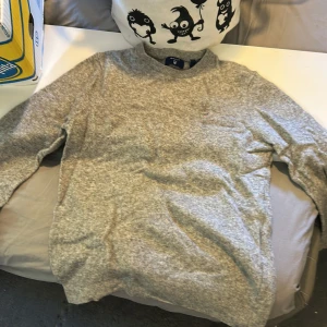 Grå stickad tröja från GANT - Mysig grå stickad tröja från GANT med rund halsringning och broderad logga på bröstet. Perfekt för lager-på-lager och passar till både jeans och chinos. Tröjan har långa ärmar och ribbade muddar för extra komfort. Passar s och xs