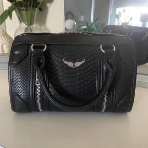 Svart handväska i skinn med ormmönster - Snygg svart handväska från Rebecca Minkoff i skinn med ormmönstrad struktur, silverfärgade detaljer och dragkedja upptill. Väskan har två handtag, avtagbar axelrem och coolt emblem framtill. Perfekt storlek för det viktigaste och ger en edgy vibe till din outfit.